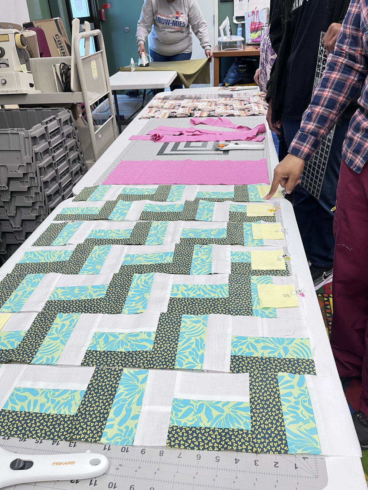 Quilt blocks in teal, yellow, and white laid out on a long studio table at the Center in the Park quilting class, with rotary cutter and rulers visible